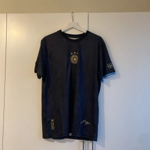 Svart fotbollströja från Adidas - Snygg svart fotbollströja från Adidas med guldiga detaljer och nummer 10 på ryggen. Tröjan har korta ärmar och en stilren design med ett emblem på bröstet. Perfekt för fotbollsfans som vill ha en unik look på planen. Special edition Messi 23/24