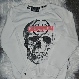 Vit tröja med dödskalletryck från Philipp Plein - Säljer en cool vit tröja från Philipp Plein med ett stort dödskalletryck och logotyp på framsidan, samt med små diamanter. Tröjan har långa ärmar och en rund halsringning. Har en grå fläck vid armen längst ner av franslim, men det märks knappt o går säkert att få bort. Pris kan diskuteras.