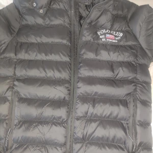 Svart jacka från Polo Club - Säljer en svart jacka  från Polo Club med dragkedja och tryck på bröstet. Jackan har en sportig look och är perfekt för kyligare dagar. Märket är broderat på framsidan och huvan har ett stort tryck , helt ny aldrig använd 
