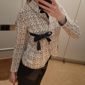 Mönstrad jacka med knytband - Snygg mönstrad jacka i grå/beige och svart med ett elegant knytband i midjan. Jackan har långa ärmar och ett unikt geometriskt mönster som ger en stilren look. Perfekt för att piffa upp vilken outfit som helst.från Odd Molly!