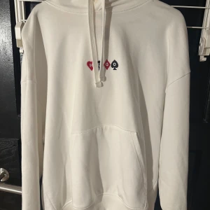 Vit hoodie från Hugo - Säljer en stilren vit hoodie från Hugo med tryck av spelkort och texten 'Full House' på baksidan. Den har en känguruficka och justerbar huva med dragsko. Perfekt för en avslappnad stil.