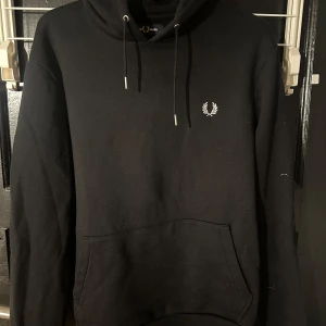 Svart hoodie från Fred Perry - Säljer en stilren svart hoodie från Fred Perry med det klassiska logotypbroderiet på bröstet. Tröjan har en känguruficka och justerbar huva med dragsko. Perfekt för en avslappnad look.