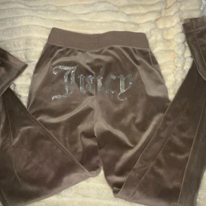 Bruna velourbyxor från Juicy Couture - Snygga bruna velourbyxor från Juicy Couture med glittrande detaljer. Byxorna har en elastisk midja och snörning framtill. Storlek  xs men jag skulle påstå att dom är typ M.