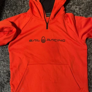 Orange hoodie från Sail Racing - Snygg orange hoodie från Sail Racing med svart logga på bröstet. Tröjan har en praktisk dragkedja vid halsen och en stor ficka framtill. Perfekt för en avslappnad stil. Köpt för 2000
