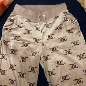 Beige mjukisbyxor från Juicy Couture - Snygga beige mjukisbyxor från Juicy Couture med ett all-over logotypmönster i brunt. Byxorna har en bekväm resårmidja med snörning och glittrande detaljer på baksidan. Har tyvärr inte taggen men köpte från JD för två år sen, o dessa är limited edition vilket e varför man inte kan hitta de på hemsidan 
