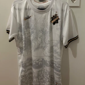 AIK STOCKHOLMS TRÖJA - Säljer en vit AIK fotbollströja från Nike med klubbens emblem på bröstet och subtilt mönster. Tröjan har korta ärmar med svarta och guldiga detaljer. Perfekt för alla AIK-fans! Produkten är helt ny och oanvänd.