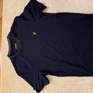 Mörkblå t-shirt från Lyle & Scott - Snygg mörkblå t-shirt från Lyle & Scott med en gul broderad logga på bröstet. Perfekt för en stilren och avslappnad look. Kortärmad och tillverkad i mjukt material för extra komfort. Storlek M. Mycket bra skick.
