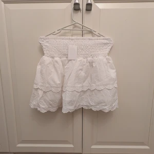 Vit kjol med volangdetaljer - Charmig vit kjol med volangdetaljer och broderade mönster. Den har en elastisk midja för bekväm passform och är perfekt för sommarens varma dagar. Kjolen är kort och har en luftig design som ger en romantisk känsla. Aldrig använd med prislapp kvar💕 perfekt till våren och sommaren💕 HELT SLUTSÅLD!