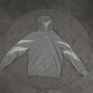 Grå hoodie från Adidas - Säljer en grå hoodie från Adidas med vita detaljer på ärmarna. Den har en klassisk huva och en liten logga på bröstet. Perfekt för en avslappnad stil.