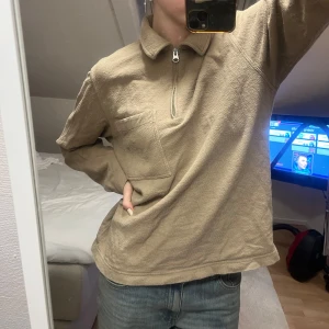 Beige tröja med dragkedja - Säljer en beige tröja med dragkedja och hög krage. Tröjan har en praktisk bröstficka och är tillverkad i ett texturerat material som ger en skön känsla. Perfekt för en avslappnad stil.