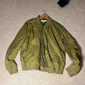 Olivgrön bomberjacka från H&M - Snygg olivgrön bomberjacka från H&M med dragkedja och ribbade muddar. Jackan har långa ärmar och praktiska fickor. Perfekt för en stilren look.