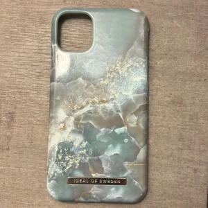 Marmorerat mobilskal från Ideal of Sweden - Snyggt mobilskal med marmorerat mönster i blå och beige toner från Ideal of Sweden. Skalet har en elegant design med en glansig yta och en diskret logotyp i guld. Perfekt för att ge din telefon ett stilrent och modernt utseende.