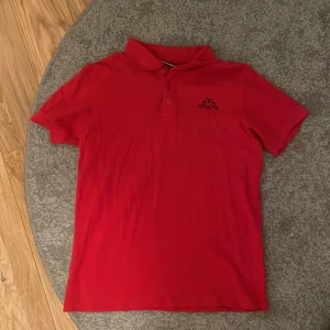 Röd pikétröja från Kappa - Säljer en snygg röd pikétröja från Kappa med klassisk krage och knappslå. Tröjan har Kappas ikoniska logga broderad på bröstet. Perfekt för en sportig och avslappnad stil.