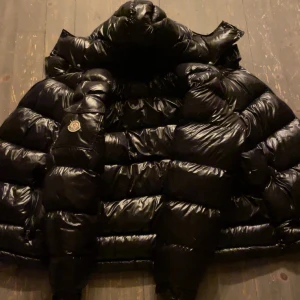 Svart dunjacka från Moncler - Snygg och varm svart dunjacka från Moncler med glansig finish. Jackan har en hög krage och Moncler-logga på ärmen. Perfekt för kyliga dagar.nytt sckick använt i ett par dagar inga konstigheter säljer pga för stor storlek säljer till ett bra pris!