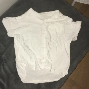Vit kortärmad skjorta från H&M - Säljer en stilren vit kortärmad skjorta från H&M. Skjortan har knappar framtill och är perfekt för en avslappnad look. Tillverkad i ett lätt och bekvämt material. Väldigt fin men lite skrynklig