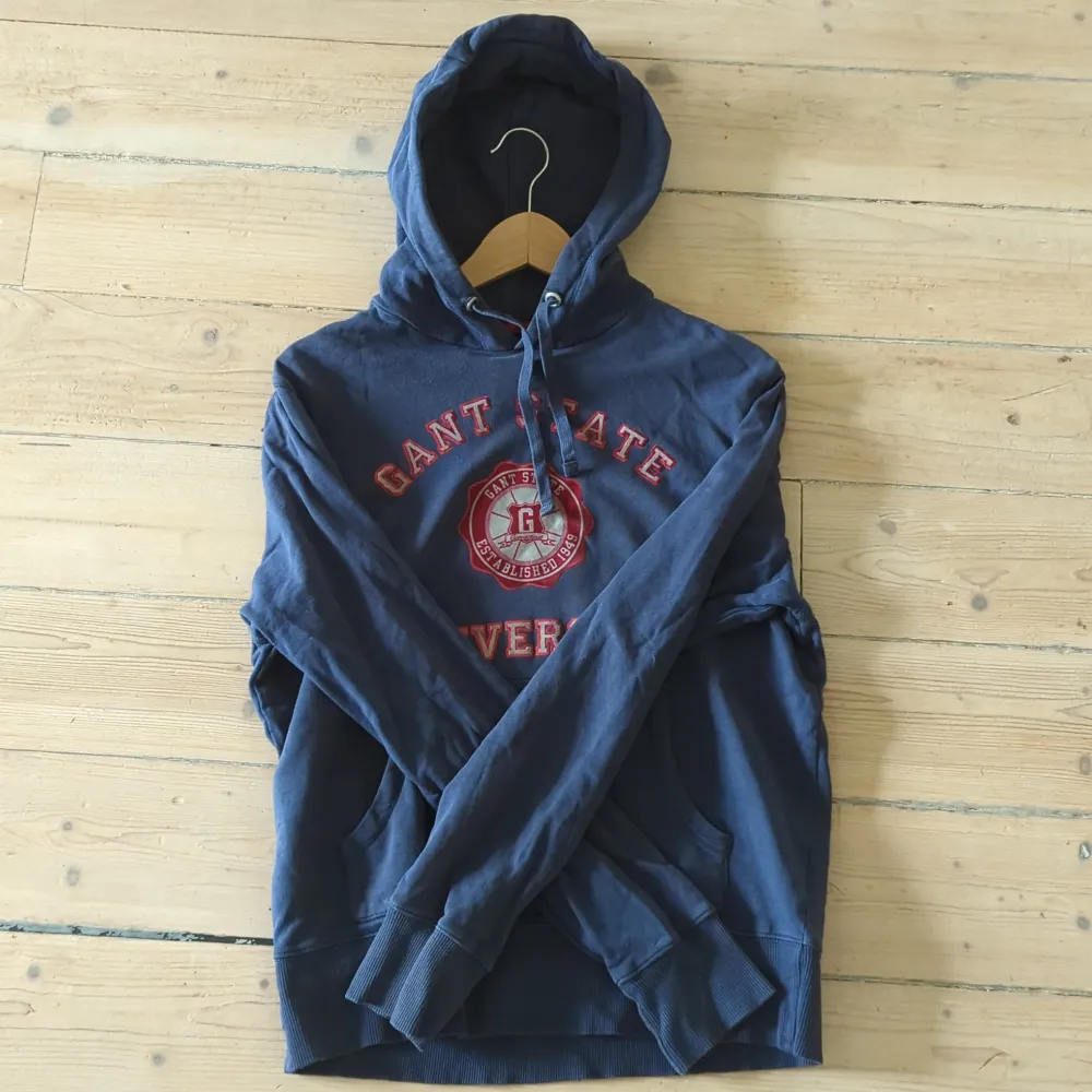 Säljer en snygg mörkblå hoodie från GANT med röd text och ett coolt universitetsmärke på bröstet. Den har en klassisk känguruficka och justerbar huva med snören. Använd några gånger men den är så gott som ny och väl omhändertagen.. Neuletakit & Villapaidat.