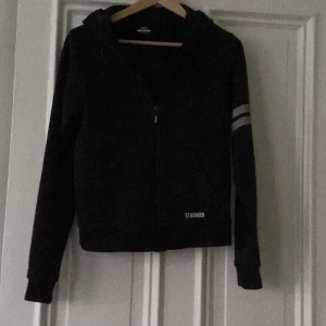 Svart jacka från Stronger M - Säljer en svart jacka från Stronger i storlek medium. Den har en dragkedja framtill och en huva. På ärmen finns det två vita ränder som ger en sportig look. Perfekt för träning eller en avslappnad dag ute.