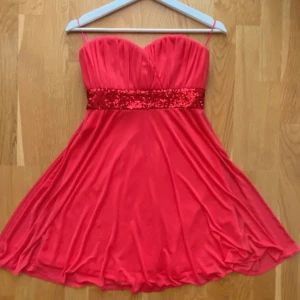 Balklänning/festklänning - Röd balklänning. Kort modell, sitter fast bra pga gummi remsa längs med kanten vid bröstet. Tråden har lossnat på vissa ställen.