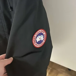 Svart vinterjacka från Canada Goose - Säljer en svart vinterjacka från Canada Goose i bra skick. Jackan har en dragkedja och knappar framtill, samt en broderad logga på ärmen. Perfekt för kalla vinterdagar med sin varma och skyddande design. Några små tecken på användning finns. Finns även ett litet håll vid sidan av jackan.