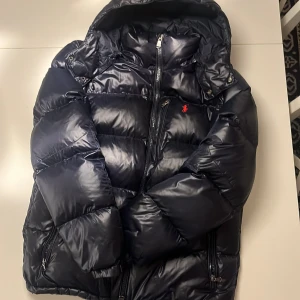 Mörkblå pufferjacka från Polo Ralph Lauren - Säljer en snygg mörkblå pufferjacka från Polo Ralph Lauren i storlek M. Jackan är perfekt för vintern med sin tjocka vaddering och har en praktisk dragkedja framtill. Den är i mycket bra skick och har en klassisk design med en liten röd logga på bröstet. Perfekt för kalla dagar! ❄️