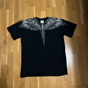 Säljer en svart t-shirt från Marcelo Burlon med ett coolt vingmönster i grått över axlarna. Perfekt för en unik och stilren look. T-shirten är kortärmad och i normal passform. Passar både till vardags och fest! Äktheten är oklar, köpte den på facebook, fråga gärna innan du köper.