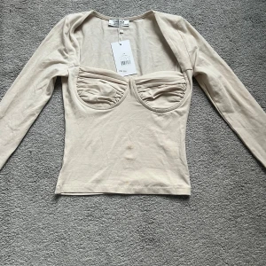 Beige topp från Chiquelle - Säljer en snygg beige topp från Chiquelle i storlek S. Toppen har långa ärmar och en unik design med rynkade detaljer vid bysten. Perfekt för en stilren look! Ny med tag kvar.