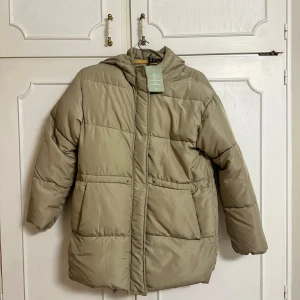 Beige dunjacka - Säljer en supermysig beige dunjacka med luva. Perfekt nu till vintern då den håller en varm. Den är i nytt skick med prislapp. Storlek XS men den är oversized