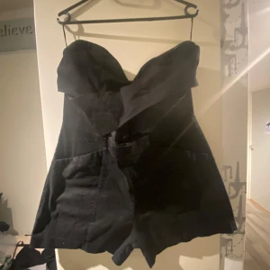 Svart axelbandslös klänning - Snygg svart axelbandslös byxdress med dragkedja i ryggen. Perfekt för festliga tillfällen. Byxdressen har en elegant och stilren design som passar bra till både höst och vinter.