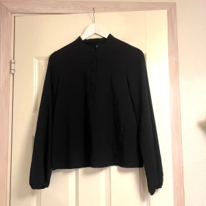 Svart långärmad topp från Vero Moda - Säljer en stilren svart långärmad topp från Vero Moda i storlek XS. Toppen har en enkel design med knappar framtill och en hög krage. Perfekt för både vardag och festliga tillfällen. Passar bra till höst- och vintergarderoben!