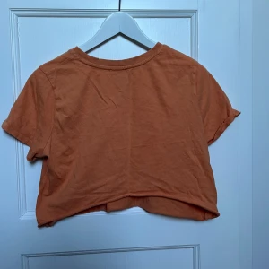 Orange croppad t-shirt - Säljer en snygg orange croppad t-shirt i bomull. Perfekt för sommardagar eller som en del av en lager-på-lager-look. Den har korta ärmar och en avslappnad passform. Superbekväm och lätt att matcha med jeans eller shorts!