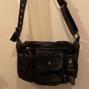 Svart axelväska i skinn - Säljer en snygg svart axelväska i skinn med flera praktiska fickor och dragkedjor. Väskan har justerbar axelrem och en cool, lite sliten look. Perfekt för vardagsbruk eller en kväll ute. 🖤