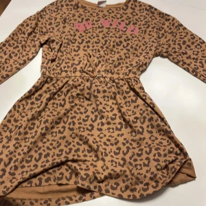 Leopardmönstrad klänning med text - Säljer en supercool leopardmönstrad klänning med texten 'BE WILD' i rosa på bröstet. Klänningen har långa ärmar och en bekväm passform, perfekt för både vardag och fest. Materialet känns mjukt och skönt mot huden. Perfekt för dig som vill sticka ut lite extra!