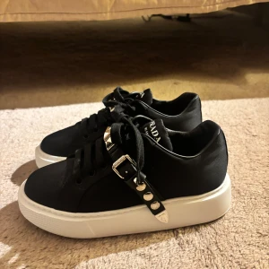 Svarta sneakers från Prada - Helt oanvända!! Snygga svarta sneakers från Prada med en chunky vit sula. De har balla nitar och ett spänne som detalj på sidan. Skorna är i syntetmaterial och har snörning framtill. Perfekta för att ge din outfit det lilla extra!