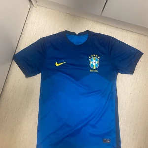 Blå Brasilien fotbollströja från Nike - Säljer en snygg blå Brasilien fotbollströja från Nike. Den har korta ärmar och det klassiska Brasilien-emblemet på bröstet. Perfekt för fotbollsfans. Tröjan är i mycket bra skick och har en cool design med Nike-loggan i gult.