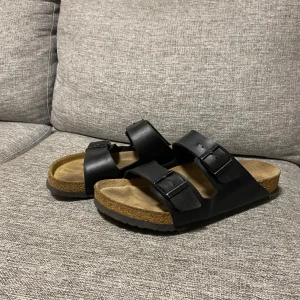 Svarta sandaler från Birkenstock - Säljer ett par klassiska svarta Birkenstock sandaler i mycket bra skick. De har två justerbara remmar och en bekväm korksula som formar sig efter foten. Perfekta för sommaren och ger en avslappnad stil. Sulan har bra grepp och är idealisk för både inomhus och utomhusbruk.