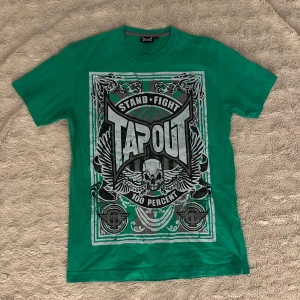 Grön T-shirt från Tapout - Säljer en grön T-shirt från Tapout med ett stort tryck på framsidan. Den har texten 'Stand Fight Tapout 100 Percent' och coola grafiska detaljer som vingar och en dödskalle. Perfekt för dig som gillar en sportig och tuff stil. T-shirten är i bomull och har en normal passform.