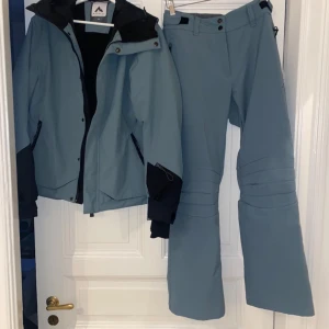 Skidjacka och skidbyxor  - Säljer ett set med blå skidjacka och matchande skidbyxor. Jackan har dragkedja och knappar, samt ficka för liftkort vid ärmsluten. Byxorna har justerbar midja och dragkedja längst ner vid benen för pjäxor. Använda men i bra skick. Det finns några få fläckar på baksidan av jackan, men de syns knappt. 