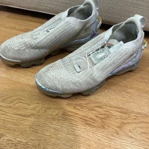 Grå Nike Vapormax sneakers - Säljer ett par grå Nike Vapormax sneakers i stickat material. De har en futuristisk design med transparent sula och elastisk snörning för en tight passform. Perfekta för både träning och vardag. Skorna är i mycket bra skick och redo för nya äventyr!