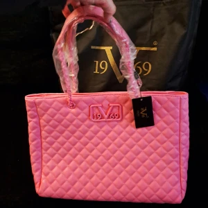 Rosa quiltad handväska från 19V69 - Säljer en superfin rosa handväska från Versace 19V69 i quiltat mönster. Väskan är i helt nytt skick (oanvänd) och har en praktisk dragkedja upptill. Perfekt för att ge en färgklick till din outfit! 💖 väskan är helt unik och köptes för 2500kr går ej att få tag på längre. 