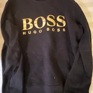 Säljer en snygg svart sweatshirt från Hugo Boss med guldig logga på bröstet. Perfekt för en stilren och avslappnad look. Tröjan har långa ärmar och är i mycket bra skick. Passar både till vardags och fest!