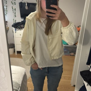 Beige vindjacka med huva - Säljer en beige vindjacka i croppad stil med huva. Jackan har långa ärmar och elastiska muddar. Perfekt för vår och sommar när det är lite kyligt ute. Den är i bra skick och passar till både jeans och klänning.