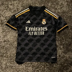 Svart Real Madrid fotbollströja från Adidas - Säljer en snygg svart Real Madrid fotbollströja från Adidas i storlek 152. Tröjan är i bra skick, använd ett fåtal gånger.