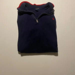 Mörkblå tröja med dragkedja från Ralph Lauren - Säljer en snygg mörkblå tröja från Ralph Lauren med en liten röd logga på bröstet. Tröjan har en dragkedja vid halsen och en röd kantdetalj. Perfekt för höst och vinter, och den är i bra skick. Passar både till vardags och lite finare tillfällen.