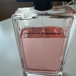 Musc Noir från Narciso Rodriguez Parfym 150ml - Säljer en elegant parfymflaska av Musc Noir från Narciso Rodriguez. Flaskan är genomskinlig med en stilren design och innehåller en rosa doftvätska. Perfekt för den som älskar en sofistikerad och feminin doft. Flaskan har en svart kork och är i utmärkt skick.