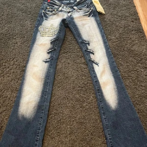 Jeans med broderad krona från Crazy Age - REDAN SÅLDA!!!                                         Snygga jeans från Crazy Age i blått med coola slitningar och en broderad krona på ena benet. De har en extrem låg midja och en bootcut passform. Midjemått:32/33 ytterbenslängden: 100 innebenslängden:94❤️