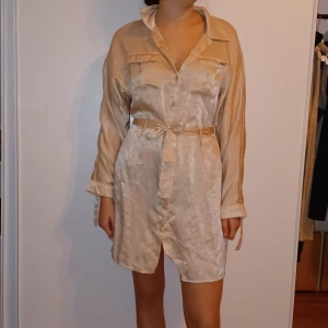 Beige sidenklänning med bälte - Snygg beige klänning i siden med lång ärm och knytbälte i midjan. Perfekt för både vardag och festliga tillfällen. Den har en klassisk krage och knappar framtill för en stilren look. Passar bra till vår och sommar.