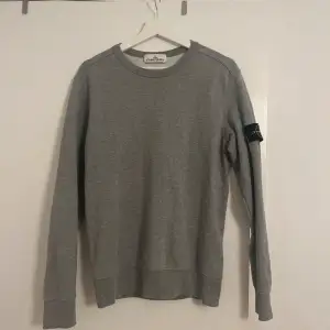 Säljer en snygg grå sweatshirt från Stone Island i mycket bra skick. Den har en klassisk rund hals och långa ärmar med det ikoniska Stone Island-märket på ärmen. Perfekt för både vardag och chill dagar. Passar till allt och är superbekväm!