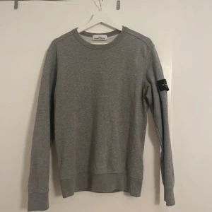 Grå sweatshirt från Stone Island - Säljer en snygg grå sweatshirt från Stone Island i mycket bra skick. Den har en klassisk rund hals och långa ärmar med det ikoniska Stone Island-märket på ärmen. Perfekt för både vardag och chill dagar. Passar till allt och är superbekväm!