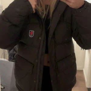  Fjällräven jacka - Säljer en grön dunjacka från Fjällräven i mycket bra skick. Jackan har en pufferstil med långa ärmar och praktiska fickor framtill. Den är perfekt för kalla vinterdagar och har en justerbar huva med gula detaljer. Supervarm och snygg för både stad och natur! Köpt förre året så den är knapt använd köpte för 6990 på länna sport