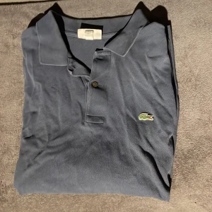 Mörkblå pikétröja från Lacoste - Säljer en klassisk mörkblå pikétröja från Lacoste i storlek M. Tröjan har en stilren design med den ikoniska krokodilloggan på bröstet och knappar vid kragen. Perfekt för både vardag och lite finare tillfällen. Skön och tidlös stil!